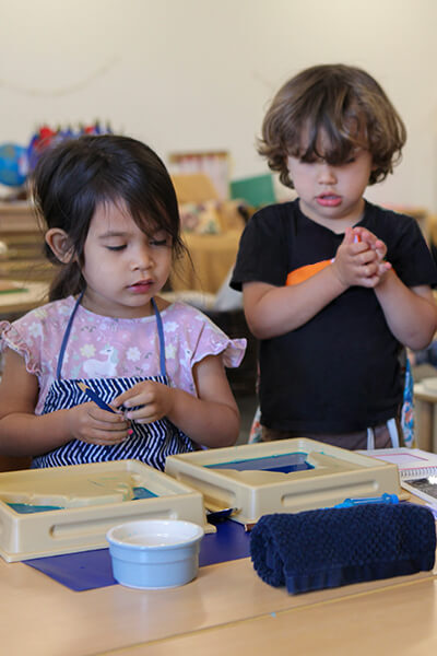 A young girl in a blue apron working with land forming Montessori tools as younger boy looks over her