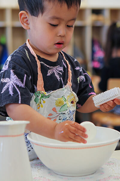 Young asian boy in blue shirt with color palm trees standing over bowl of water learning to wash their hands with a white brush