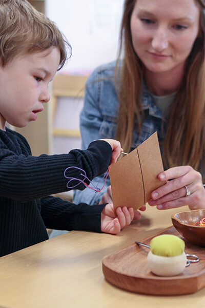 Young boy learning to sew using brown cardstock with assistance of guide