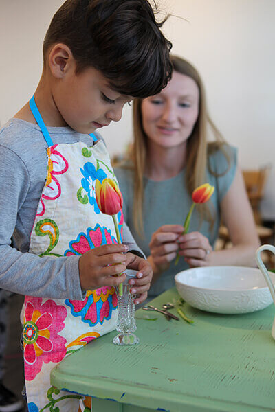 Young boy learning how to create a flower centerpiece with orange and yellow tulips in a clear vase assisted by a guide