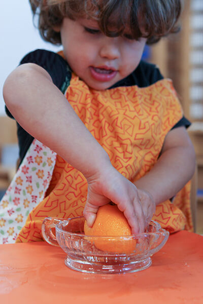 A seated young boy wearing a orange apron, holding a orange and squeezing the juice out of it