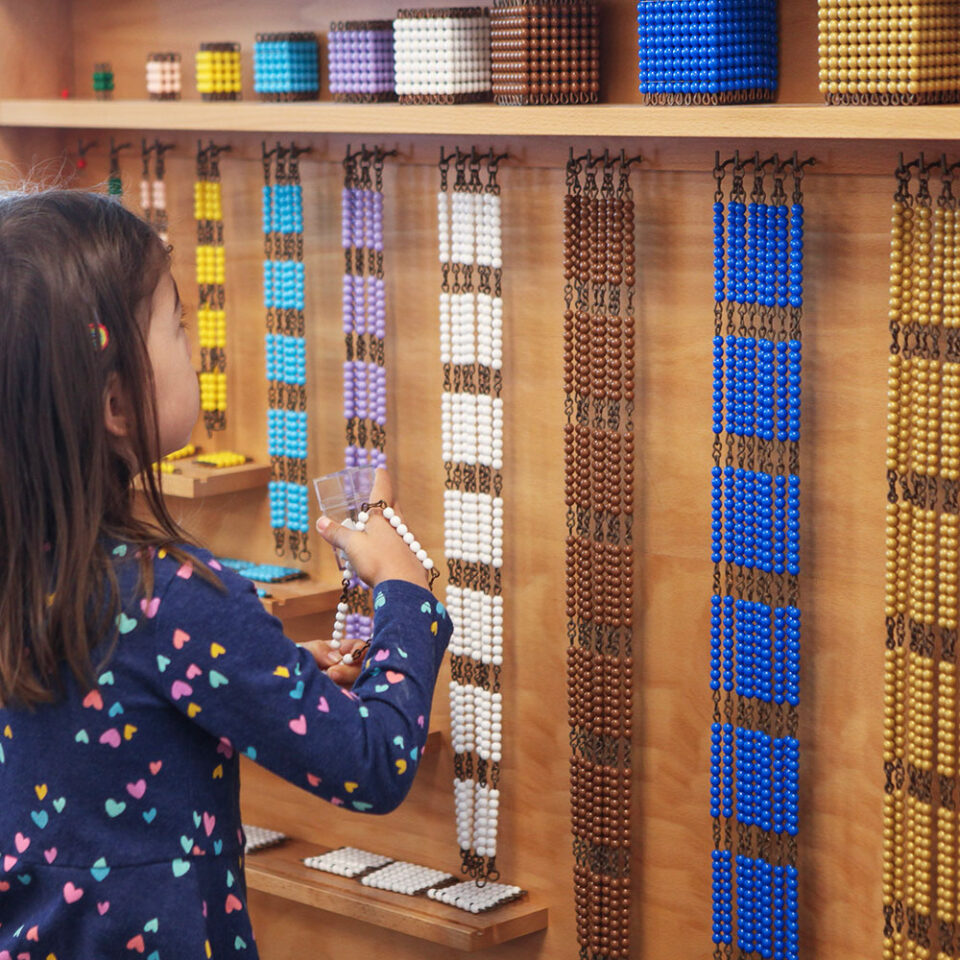 Young girl in blue dress covered with colorful hearts standing in front of wall of Montessori Golden beads
