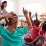 Group of young kids seated raising their hands as teacher is reading to them