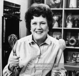 Black and white photograph of Julia Child in a kitchen setting holding a metal whisk in her right hand and ladle in the left