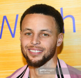 Head shot of Steph Curry smiling and wearing a pink jacket against a yellow background