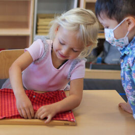Seated young blond girl buttoning a red shirt on a Montessori practical life teaching tool as a young asian boy wearing a surgical masks looks over her