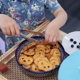 Close up on young boy in a blue Hawaiian inspired shirt holding a pair of small metal tongs picking up pretzels
